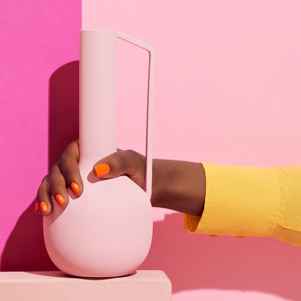 Hand mit leuchtend korall-orangen manikürten Nägeln hält weiße Keramikvase vor rosa und gelbem farblich abgesetztem Hintergrund und präsentiert Manucurist Sunset Nagellack