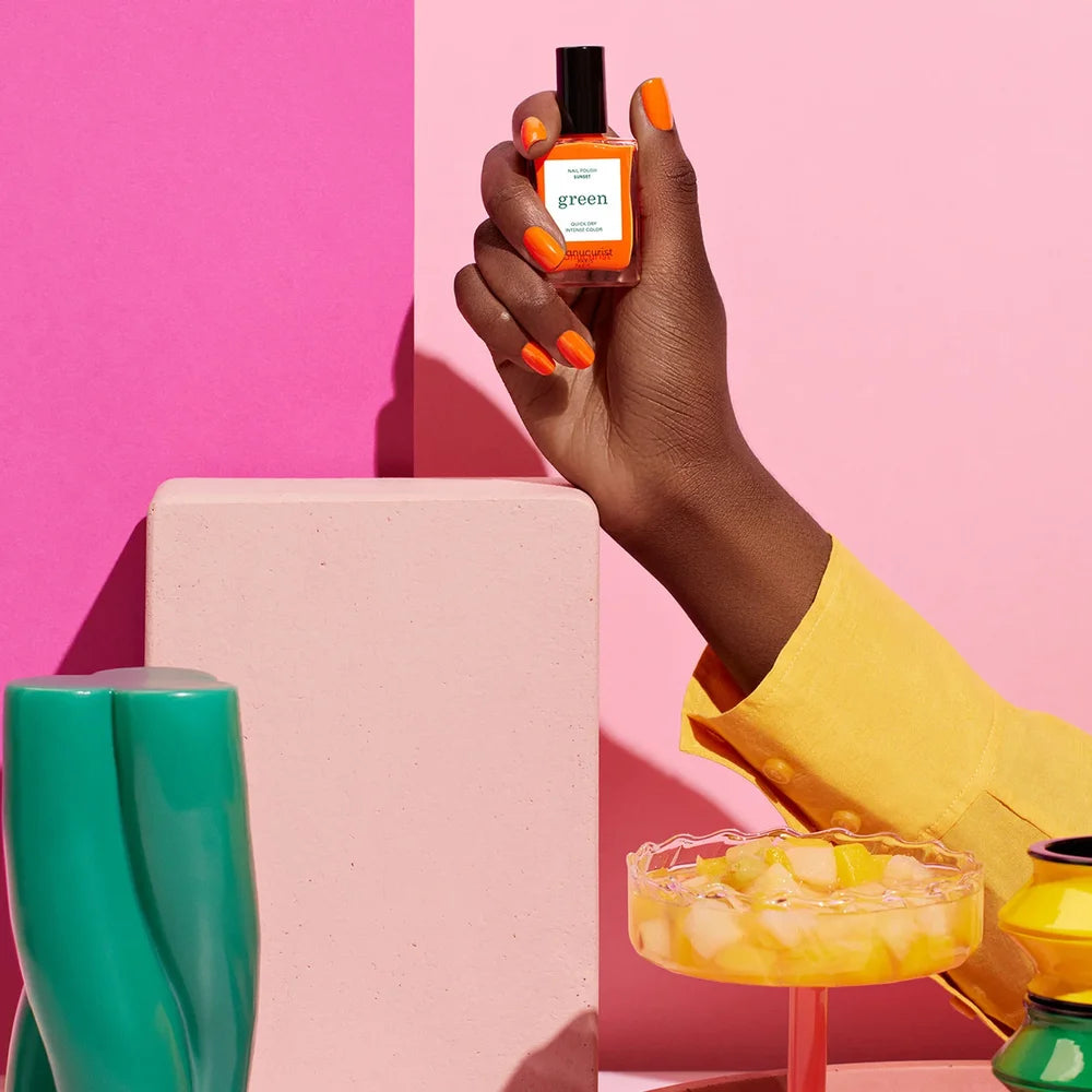 Hand mit korall-orangen manikürten Nägeln hält Manucurist Paris Green Nagellack in Sunset vor leuchtend rosa Hintergrund mit buntem Cocktailglas