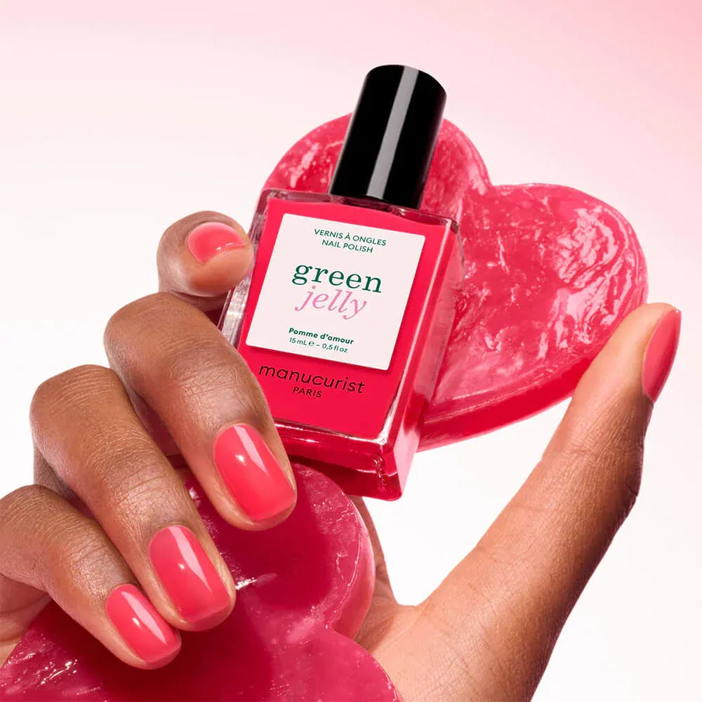 Hand hält Manucurist Green Jelly Nagellack Pomme d’Amour in leuchtendem Rot-Pink neben roten Herzen