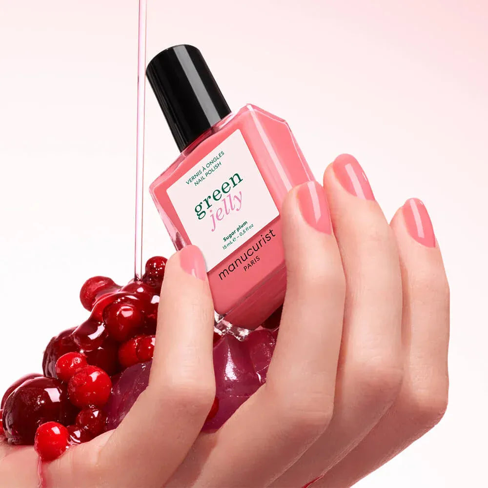 Hand hält Manucurist Green Jelly Nagellack Sugar Plum in fruchtigem Pink neben roten Kirschen