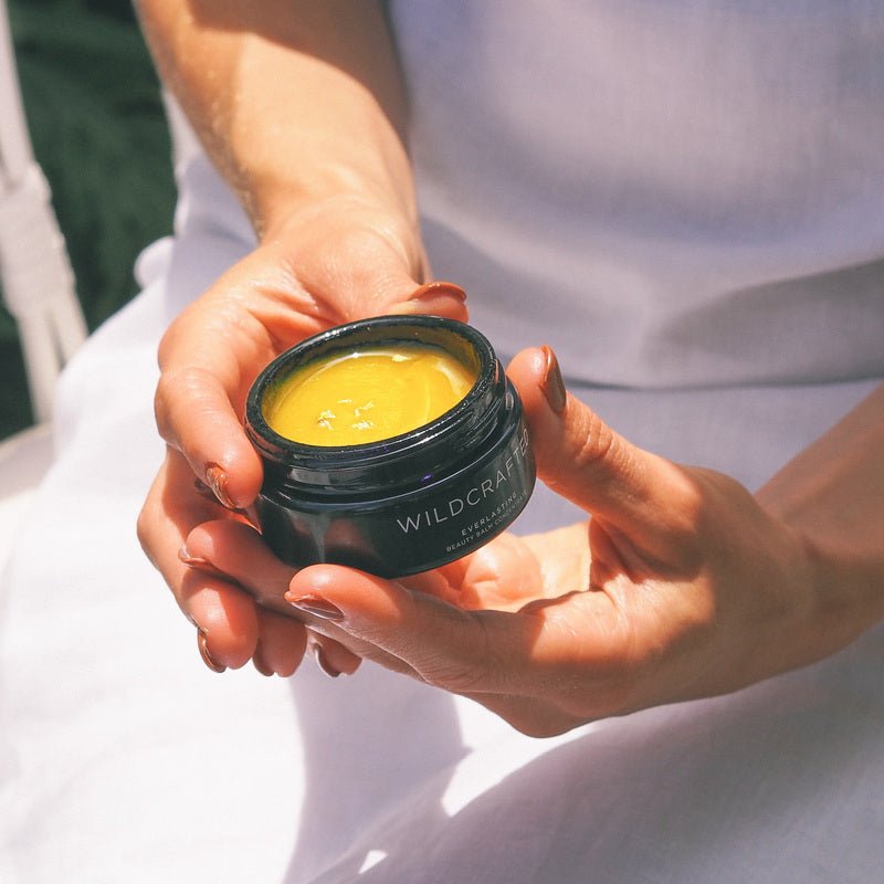 Hände halten geöffneten Wildcrafted Organics Everlasting Beauty Balm Tiegel mit goldgelber Balm-Textur